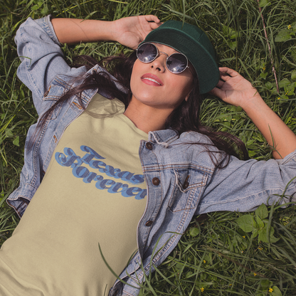 Woman lying in green grass wearing a light tan t-shirt that says “Texas Forever” in blue retro font. She has on round sunglasses, a green cap, and a denim jacket, looking relaxed and confident. Bold white text on the image reads, “Raised Right. Resting Easy.” with the Lone Star Roots logo in the top left.