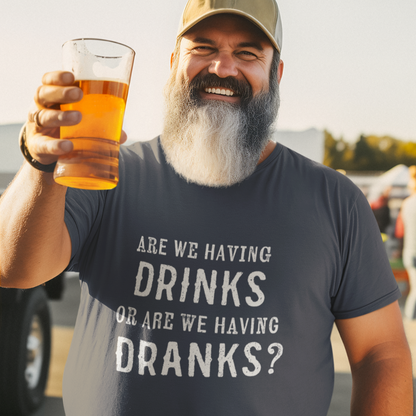 Man holding a beer and smiling while wearing an Are We Having Drinks or Dranks t-shirt at an outdoor event.
