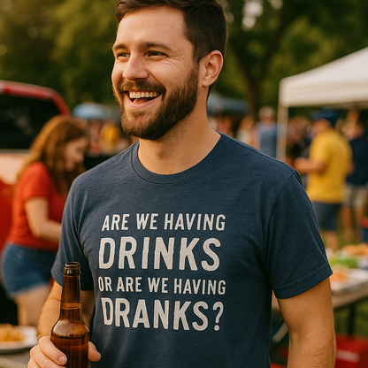 Man holding a beer and smiling while wearing an Are We Having Drinks or Dranks t-shirt at an outdoor event.